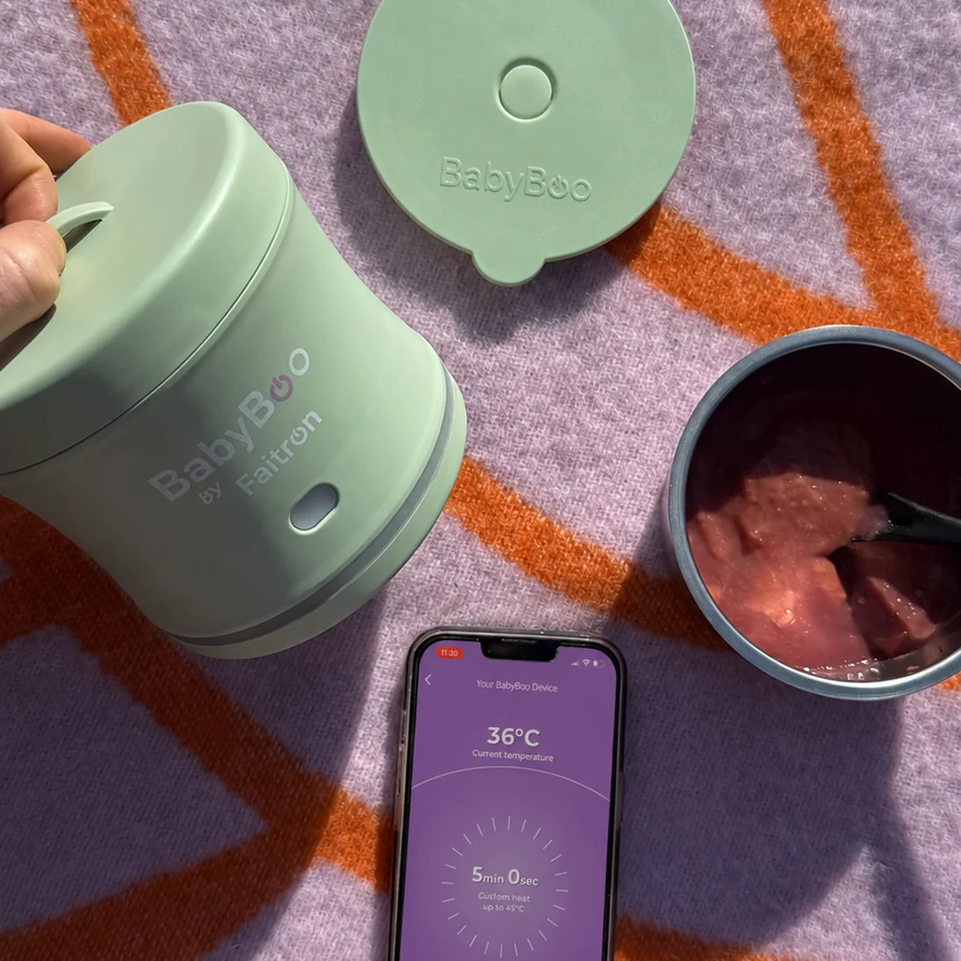 Top-down outdoor flat-lay on a lilac and orange picnic blanket — sage-green BabyBoo Food Warmer held in a hand with the matching lid resting beside it, an iPhone displaying the BabyBoo app at 36°C with a 5-minute timer, and the open 304 stainless steel inner dish filled with a warm purée meal