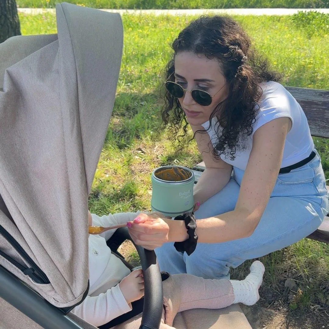 Mother in jeans and a white t-shirt sitting on a park bench in a green field, holding the sage-green BabyBoo Food Warmer with the open inner dish toward her baby in a stroller, the baby's hand reaching for the spoon